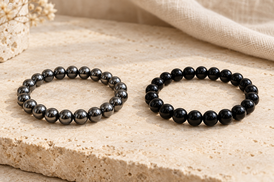  Metallic gray hematite bracelet beside a polished black obsidian bracelet on a beige stone surface.