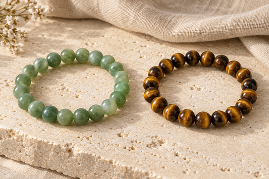 Green jade and golden-brown tiger’s eye bead bracelets displayed side by side on a beige stone surface.